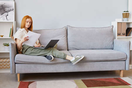 Full length portrait of adult red haired woman using laptop on sofa while working from home, copy spaceの写真素材