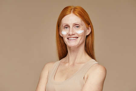 Minimal portrait of freckled red haired woman using face cream and smiling at camera, copy spaceの写真素材