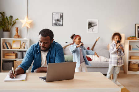 Portrait of African-American man using laptop while working at home with children playing in...