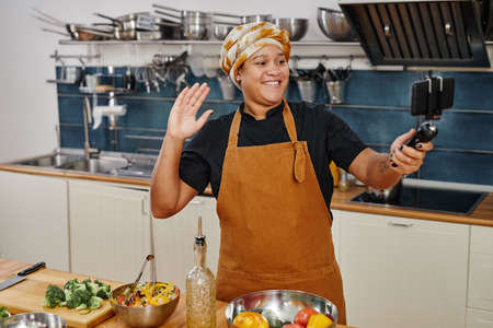 Portrait of smiling woman holding smartphone while recording videos in kitchen, copy spaceの写真素材