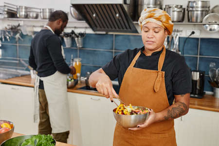 Waist up portrait of tattooed woman mixing vegetable salad while working in professional kitchen,...