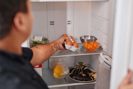 Back view portrait of unrecognizable woman taking fresh eggs out of refrigerator in kitchen, copy...