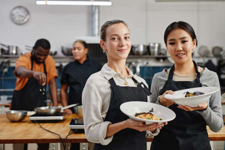 Waist up portrait of two young women wearing aprons and holding gourmet dishes in professional kitchen, copy spaceの写真素材