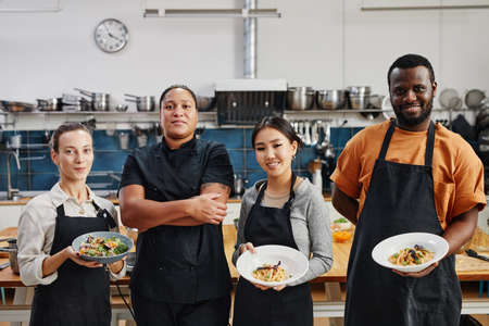 Diverse group of people wearing aprons and holding gourmet dishes in professional kitchenの写真素材