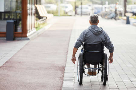 Rear view of mature man with disability riding in wheelchair along the streetの写真素材