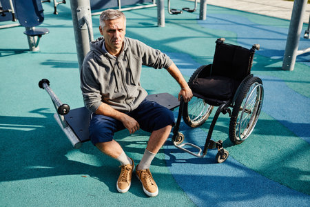 Portrait of mature man looking at camera while sitting on the bench near the wheelchair he recovering at outdoor gymの写真素材