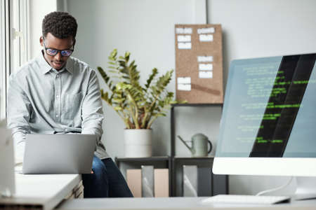 Portrait of young African-American man using laptop while sitting by window in software development office, code screen in foreground, copy spaceの写真素材
