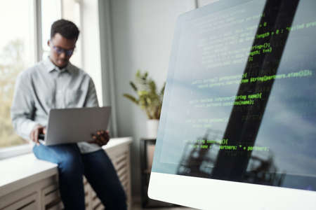 Blurred portrait of young African-American man using laptop while sitting by window in software development office, code screen in foreground, copy spaceの写真素材
