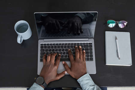 Minimal top view of unrecognizable African-American man using laptop while working at black desk background, focus on hands at keyboard, copy spaceの写真素材