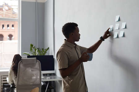 Side view portrait of young African-American man placing sticker notes on wall while planning project in office, copy spaceの写真素材