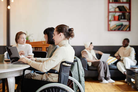 Student sitting in wheelchair and using laptop in his online study with his classmates at the tableの写真素材
