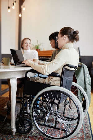 Student with disability sitting in wheelchair and studying on laptop while sitting at the table with his classmatesの写真素材