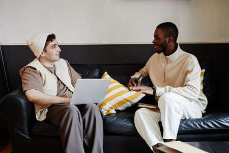 Two friends sitting on sofa with books and laptop and talking, they preparing for exams togetherの写真素材