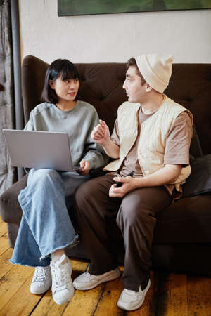 Two students sitting on sofa using laptop and discussing online education together in the roomの写真素材