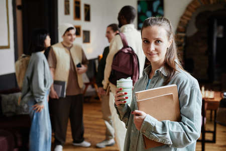 Portrait of young woman holding textbooks and coffee drink smiling at camera while studying in the universityの写真素材