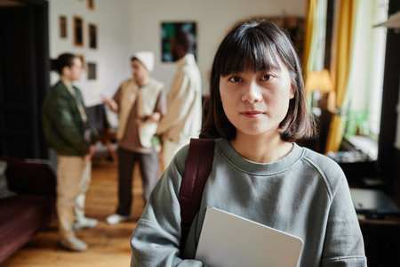 Portrait of Asian student with backpack and textbook looking at camera standing in the classroom with students in the backgroundの写真素材