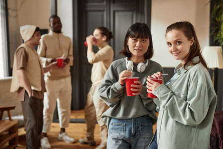 Portrait of young women standing with drinks and smiling at camera while visiting the party with their friends in the backgroundの写真素材