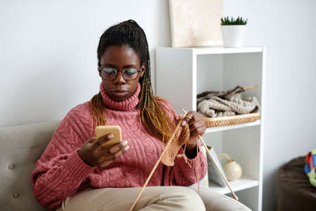Portrait of young African-American woman using smartphone while knitting at home and enjoying cozy weekend, copy spaceの写真素材