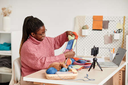 Portrait of smiling African-American woman knitting at home and recording video or livestream, copy spaceの写真素材