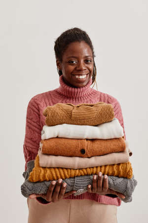 Minimal waist up portrait of young African-American woman holding stack of hand knit sweaters and smiling at cameraの写真素材