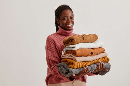Waist up portrait of young African-American woman holding stack of hand knit sweaters and smiling at camera, copy spaceの写真素材