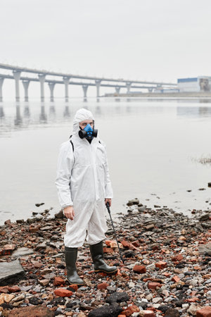 Full length portrait of male worker wearing hazmat suit by water outdoors, industrial waste concept, copy spaceの写真素材
