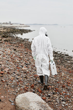 Vertical back view portrait of worker wearing hazmat suit walking by water outdoors and carrying samples case, industrial waste conceptの写真素材
