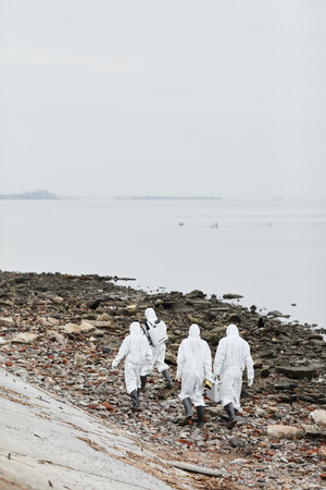 Vertical wide angle view at group of workers wearing hazmat suits walking by water outdoors , toxic waste concept, copy spaceの写真素材