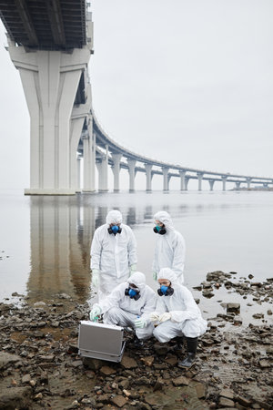 Vertical full length view view at group of people wearing hazmat suits collecting probes by water, toxic waste and pollution conceptの写真素材