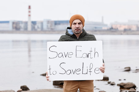 Portrait of man holding Save Earth sign and looking at camera while protesting at environmental disaster site, copy spaceの写真素材