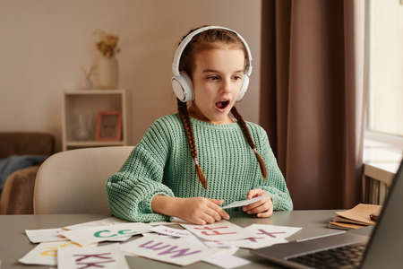 Little girl in wireless headphones sitting at the table and learning to pronounce new english words during online lesson on laptopの写真素材