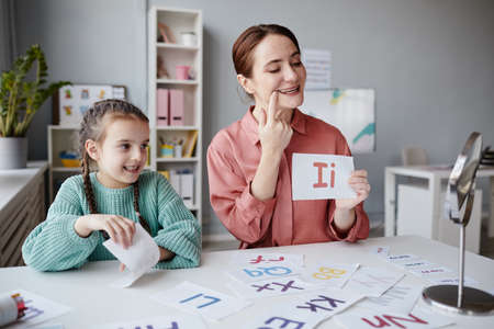 Tutor teaching to read the English letter in right way, while she sitting at the table together with girl, they speaking and looking at mirrorの写真素材