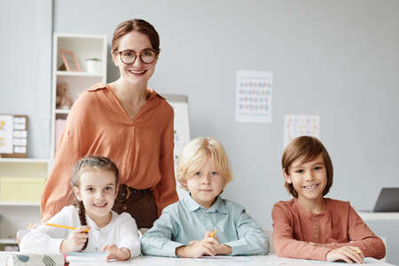 Portrait of young teacher in eyeglasses smiling at camera with group of children sitting at the table in the classの写真素材