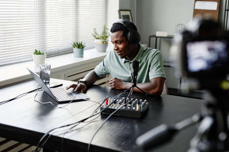 Portrait of young African-American man recording podcast in studio and using laptop