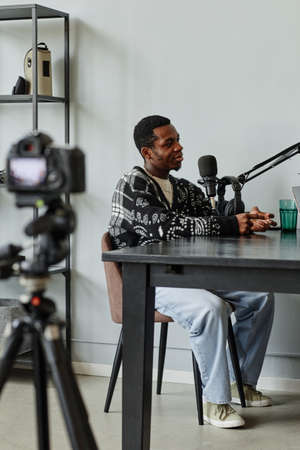 Vertical full length portrait of African-American creator speaking to microphone while recording podcast in studioの写真素材