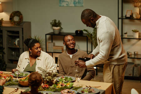 Side view portrait of African-American man pouring wine while enjoying Christmas dinner with family at homeの写真素材