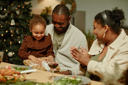 Portrait of cute African-American girl opening present while enjoying Christmas with familyの写真素材