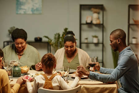Portrait of modern African-American family holding hands while saying grace during dinner party at homeの写真素材