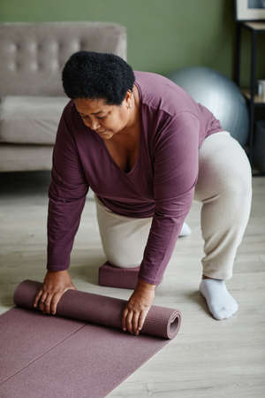 Vertical full length portrait of black senior woman unrolling yoga mat for home workoutの写真素材