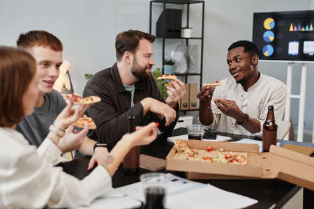 Business colleagues eating pizza at the table and talking to each other during lunch at officeの写真素材