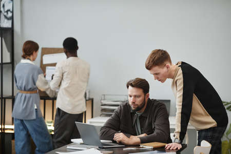 Designer sitting at his workplace and working on laptop, he discussing something with his colleague...