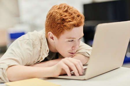 Portrait of red haired teenage boy laying head on desk while using laptop in school, copy spaceの写真素材