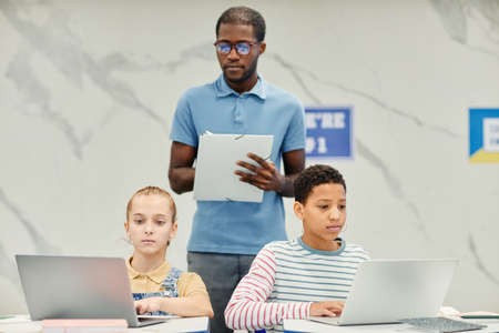 Portrait of two teenage girls using laptops in modern school classroom with African-American teacher watching them work, copy spaceの写真素材
