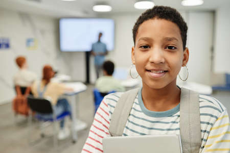 Portrait of smiling teenage girl holding books and looking at camera in school classroom, copy spaceの写真素材