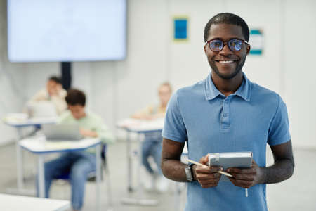 Waist up portrait of young African-American teacher smiling at camera with children in background, copy spaceの写真素材