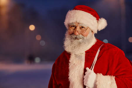 Horizontal medium close-up portrait of cheerful Santa Claus wearing white and red outfit with eyeglasses standing outdoors in evening looking at cameraの写真素材