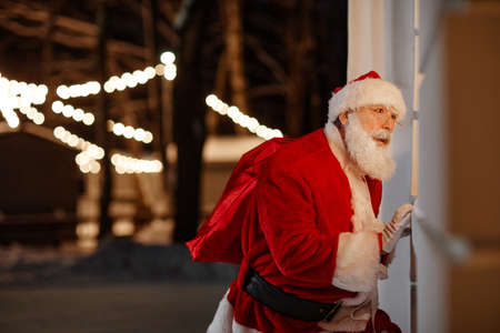 Portrait of Santa Claus delivering gifts standing outdoors peeping through window checking if family sleepingの写真素材