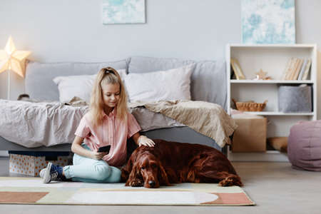 Full length portrait of cute blonde girl playing with pet dog on floor in cozy home interior , copy spaceの写真素材