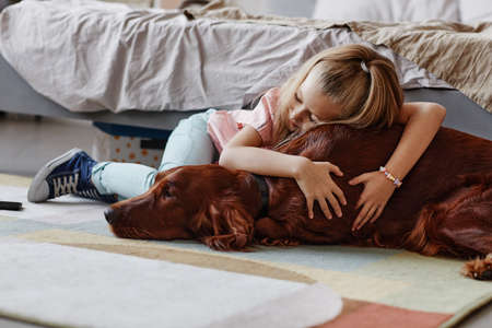 Full length portrait of cute blonde girl embracing pet dog on floor in cozy home interiorの写真素材