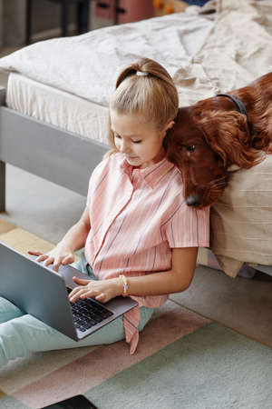 Vertical portrait of cute blonde girl using laptop while sitting on floor at home with pet dog